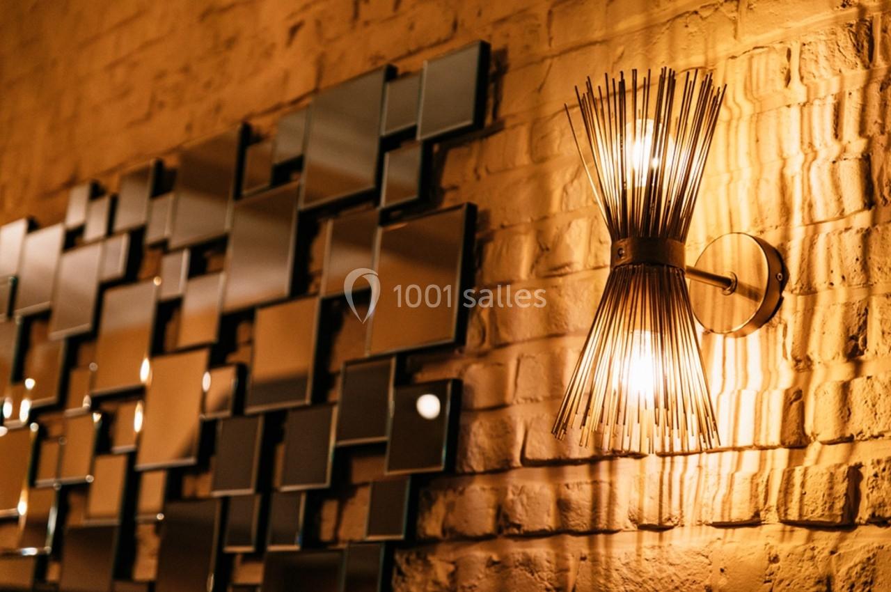 Applique murale dorée allumée sur un mur en briques, près d'un assemblage de miroirs géométriques.