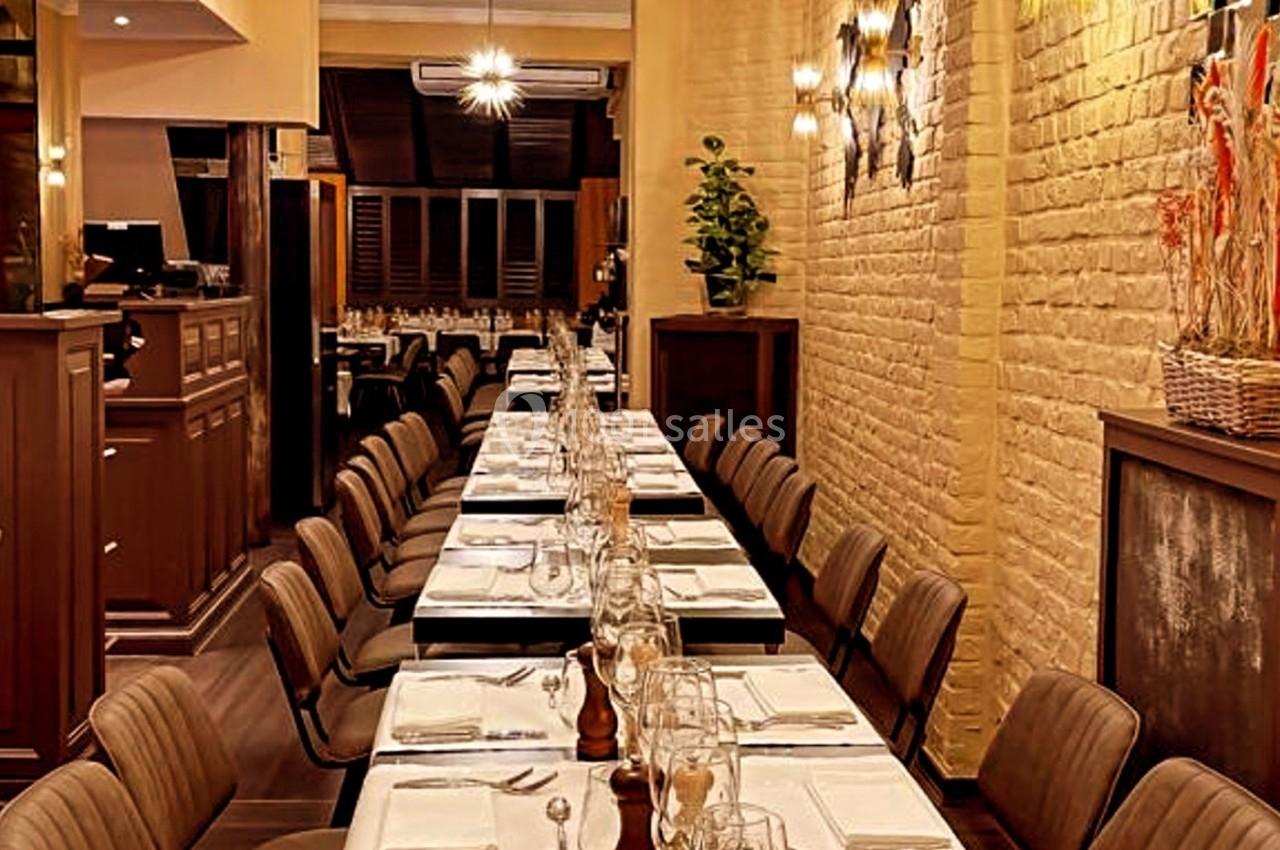 Salle de restaurant avec une longue table dressée, chaises alignées, éclairage chaleureux et murs en briques claires.
