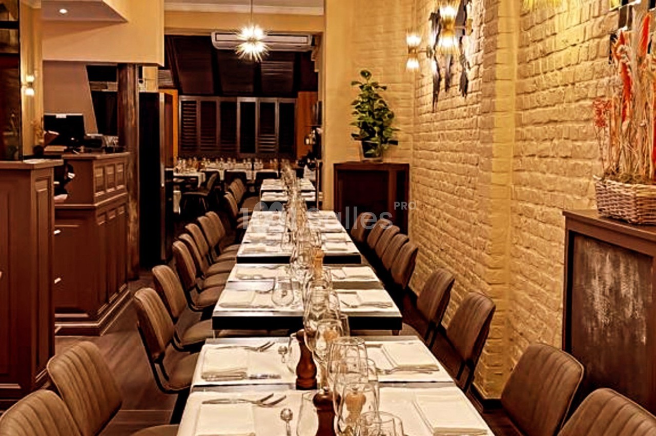 Salle de restaurant avec une longue table dressée, chaises alignées, éclairage chaleureux et murs en briques claires.