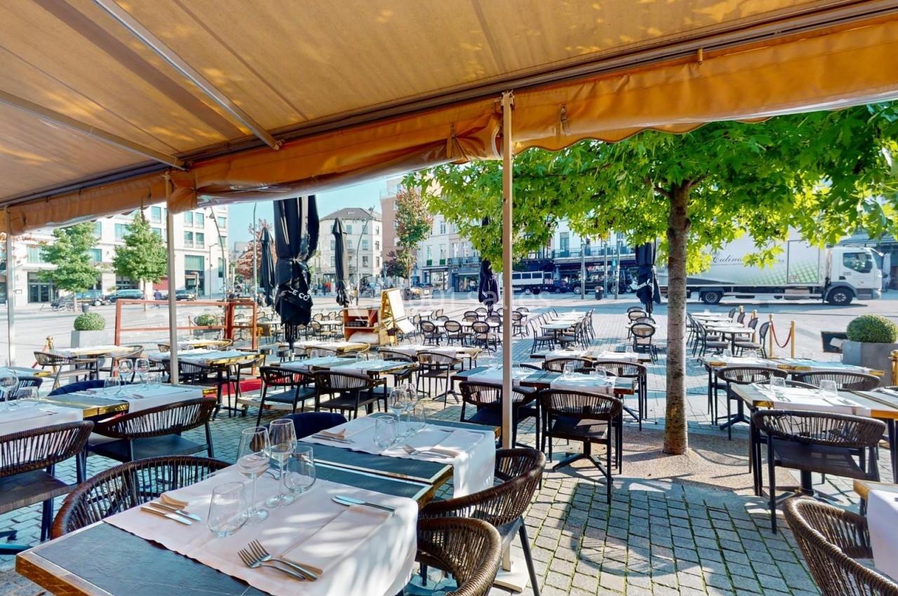 Terrasse de restaurant avec tables dressées sous un auvent, donnant sur une place arborée et des bâtiments en arrière-plan.