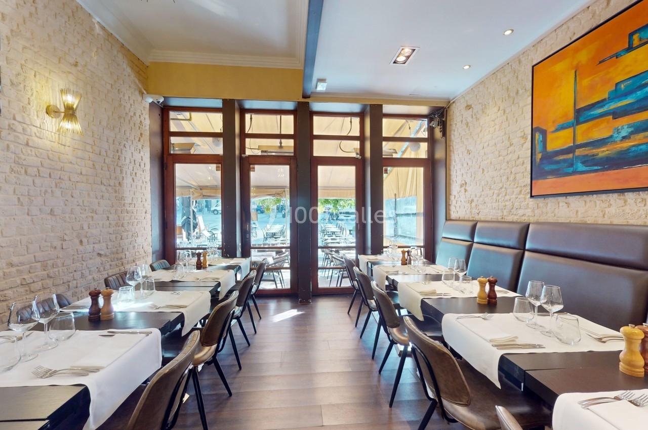 Salle de restaurant lumineuse avec tables dressées, murs en pierre, grandes baies vitrées et tableau coloré au mur.