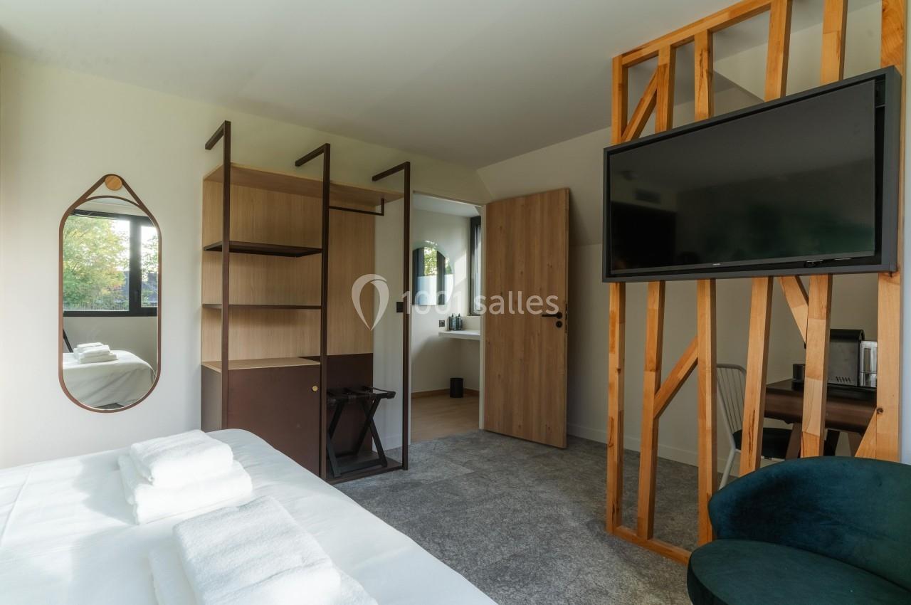 Chambre lumineuse avec lit double, étagères ouvertes, télévision murale et grande fenêtre donnant sur un espace verdoyant.