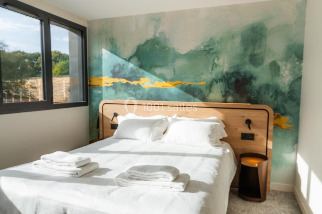 Chambre lumineuse avec un lit double, des draps blancs, une tête de lit en bois et un mur décoré d'une fresque artistique.