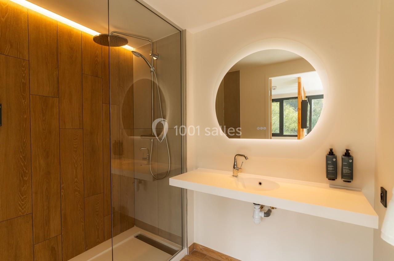 Salle de bain moderne avec douche à l'italienne, mur en bois, miroir rond éclairé et lavabo blanc.