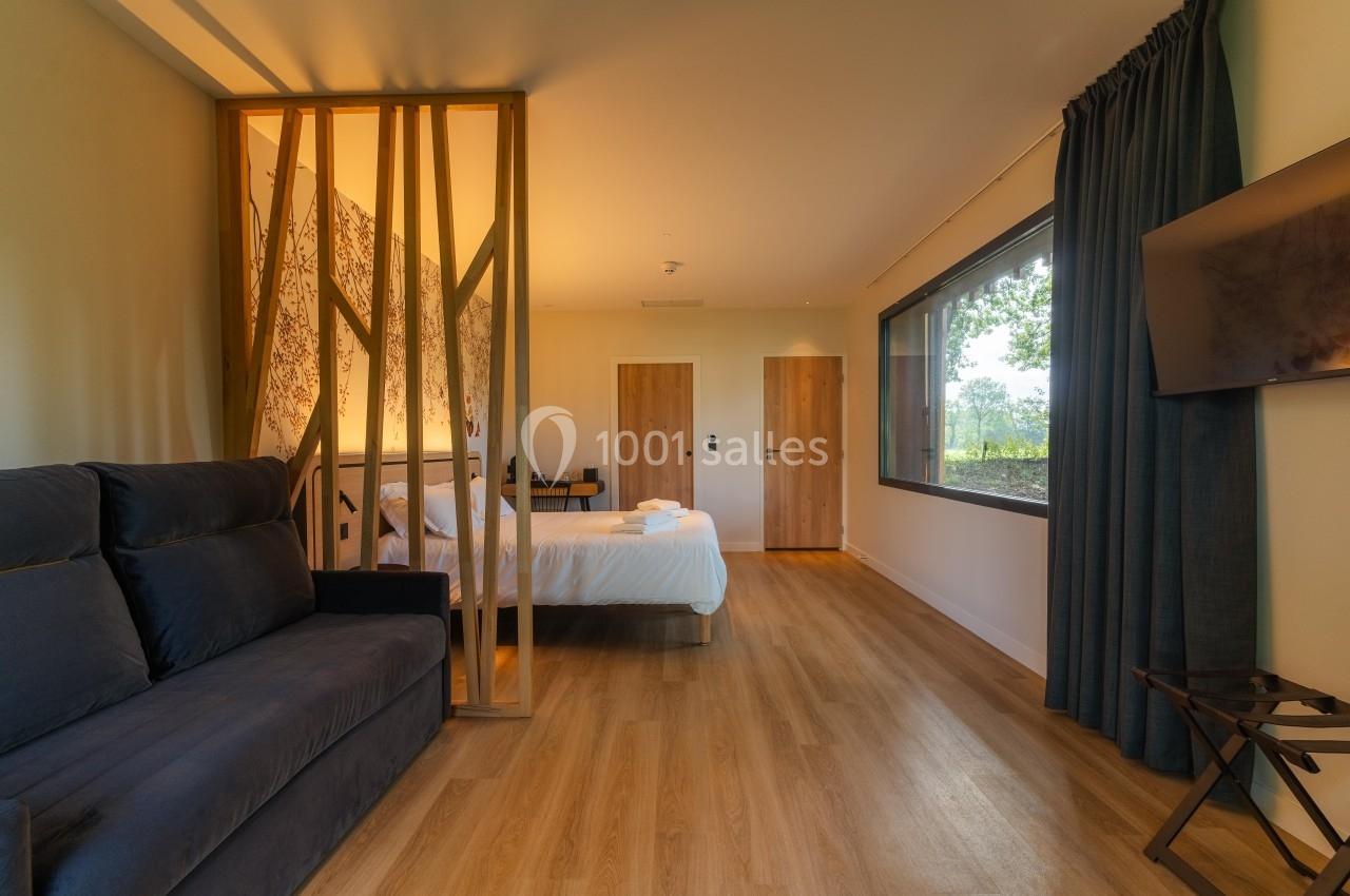 Chambre lumineuse avec lit double, canapé, cloison en bois, grande fenêtre et sol en parquet.