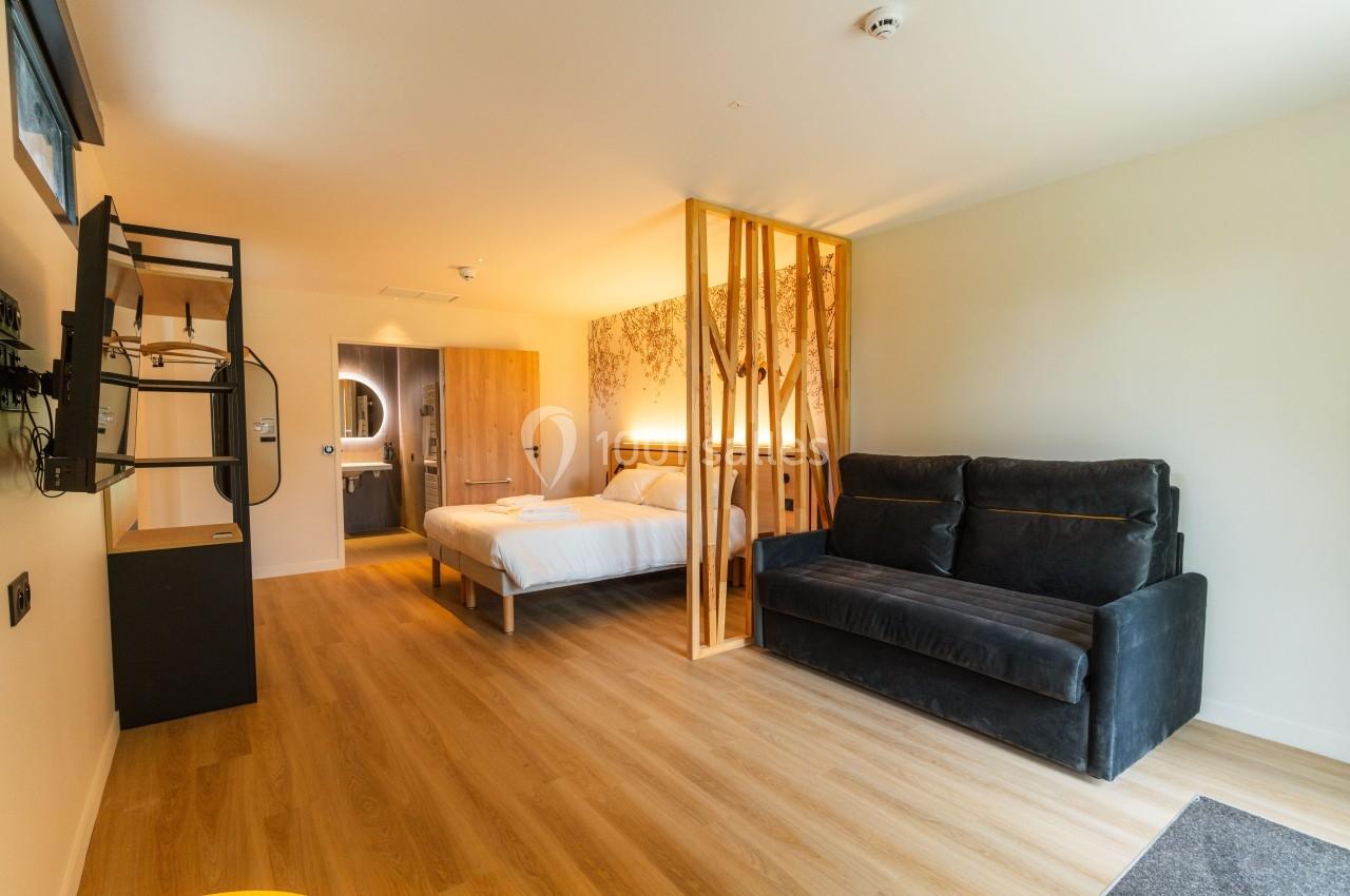 Chambre moderne avec lit double, canapé, cloison en bois, télévision murale et salle de bain visible en arrière-plan.