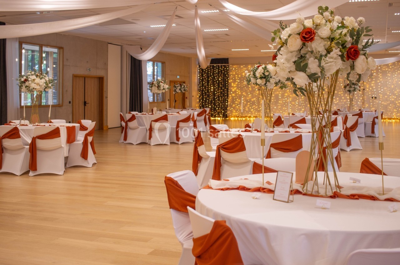 Salle de réception décorée avec des tables rondes, nappes blanches, chaises ornées de rubans orange et arrangements floraux.