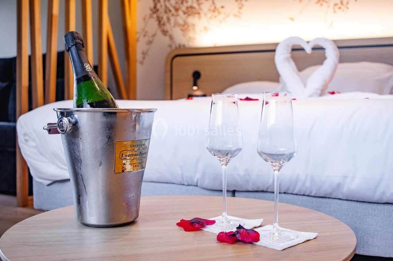 Seau à champagne, deux flûtes et pétales de rose sur une table devant un lit décoré avec des serviettes en forme de cœur.