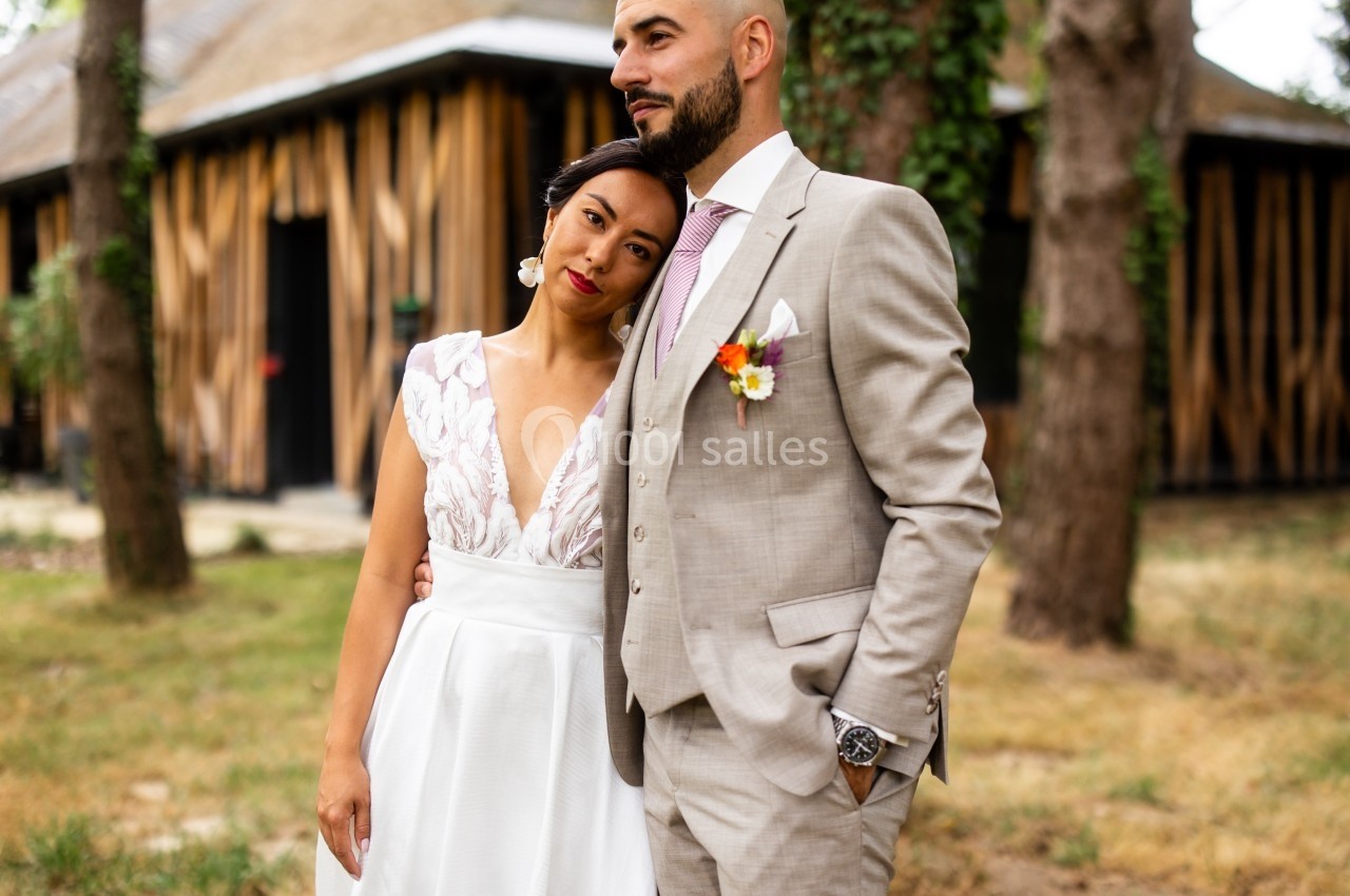 Une mariée en robe blanche repose sa tête sur l'épaule du marié en costume beige, devant un bâtiment en bois.