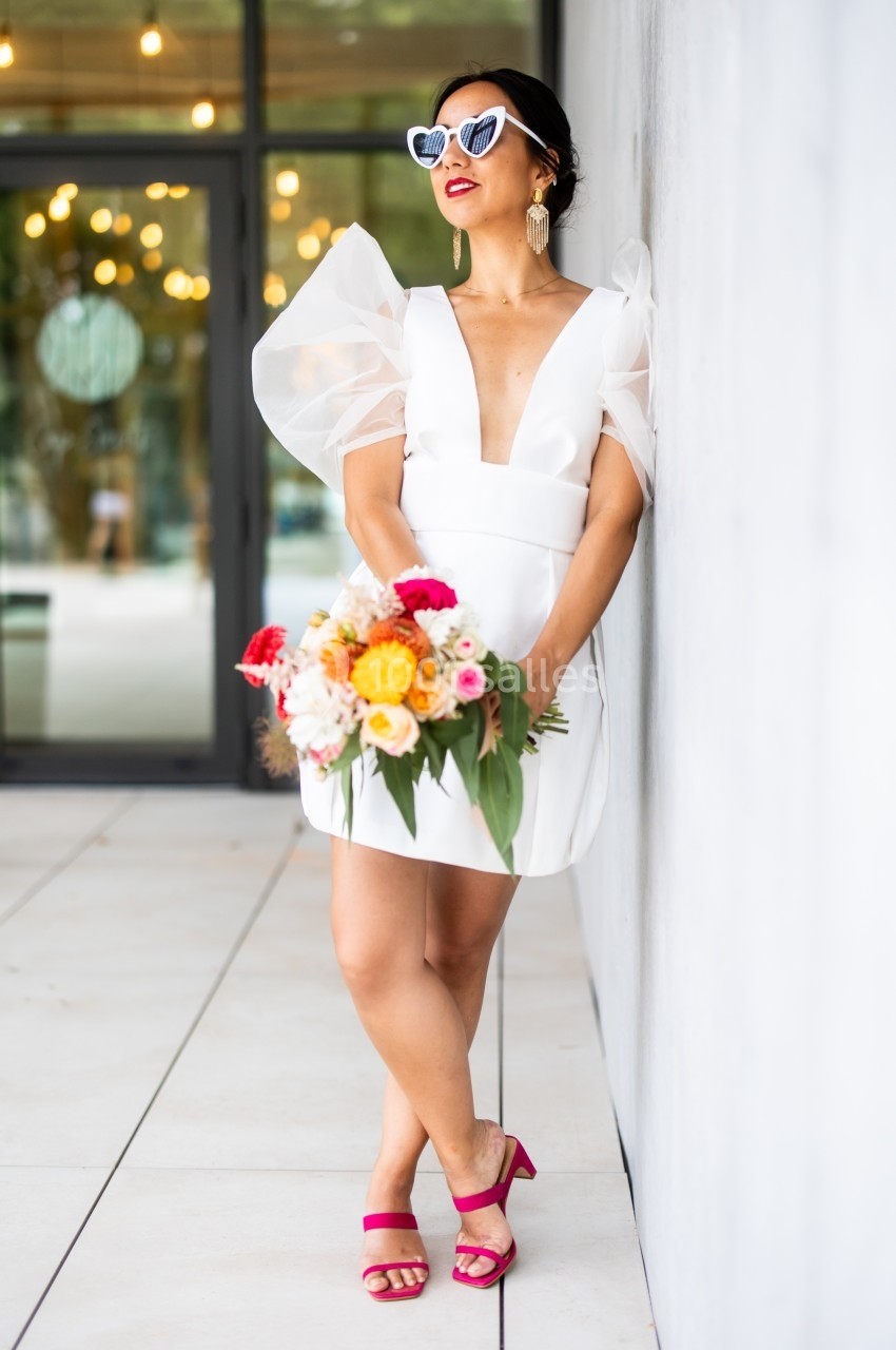 Femme en robe blanche élégante tenant un bouquet de fleurs colorées, appuyée contre un mur clair.
