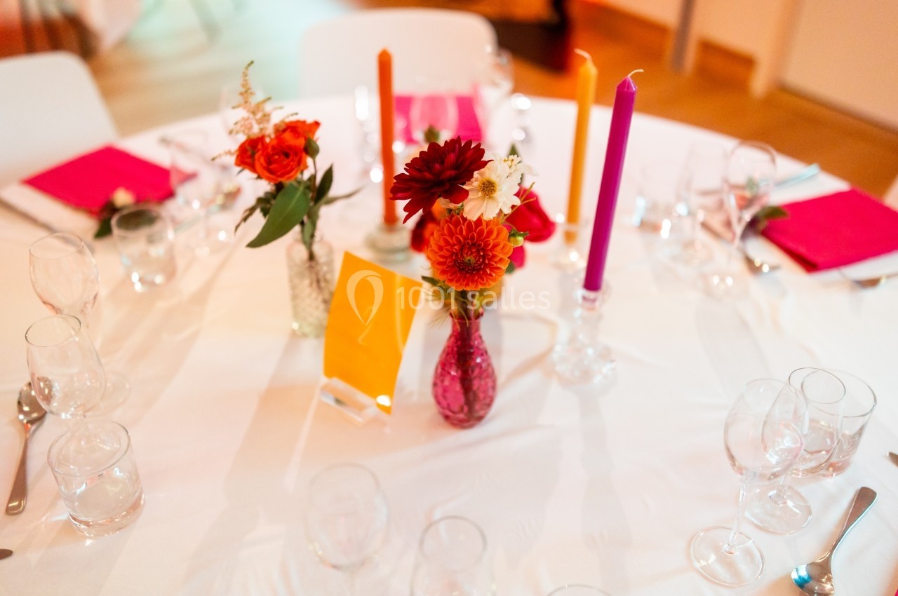 Table décorée avec des fleurs colorées, bougies orange et violettes, et serviettes roses sur une nappe blanche.