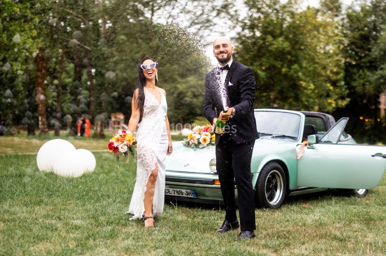 Un couple en tenue de mariage célèbre avec une bouteille de champagne devant une voiture décapotable verte.