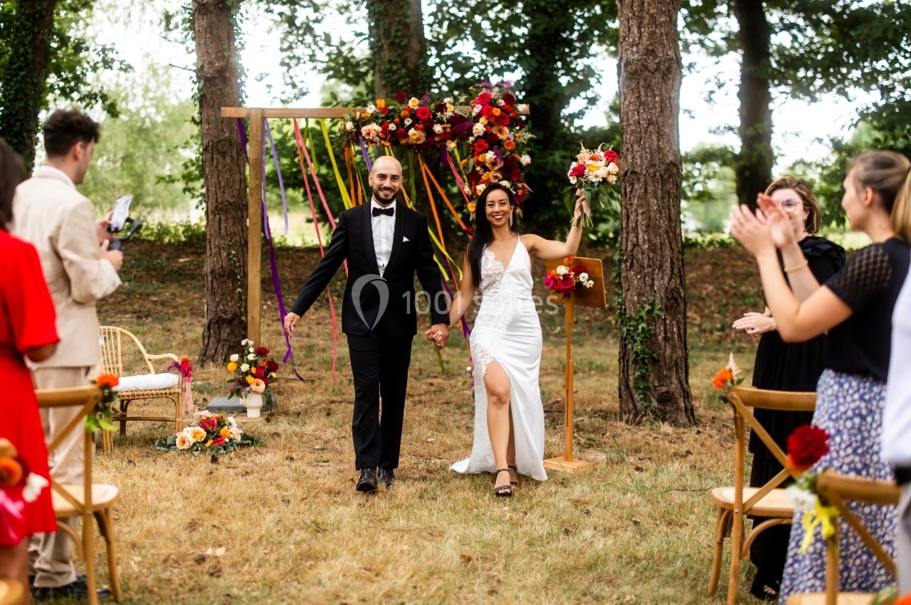 Un couple souriant marche main dans la main après une cérémonie de mariage en plein air, entouré d'invités applaudissant.