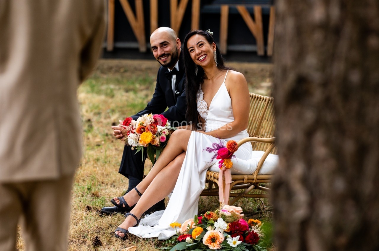 Un couple assis sur une chaise en rotin, souriant, entouré de fleurs colorées dans un cadre extérieur.