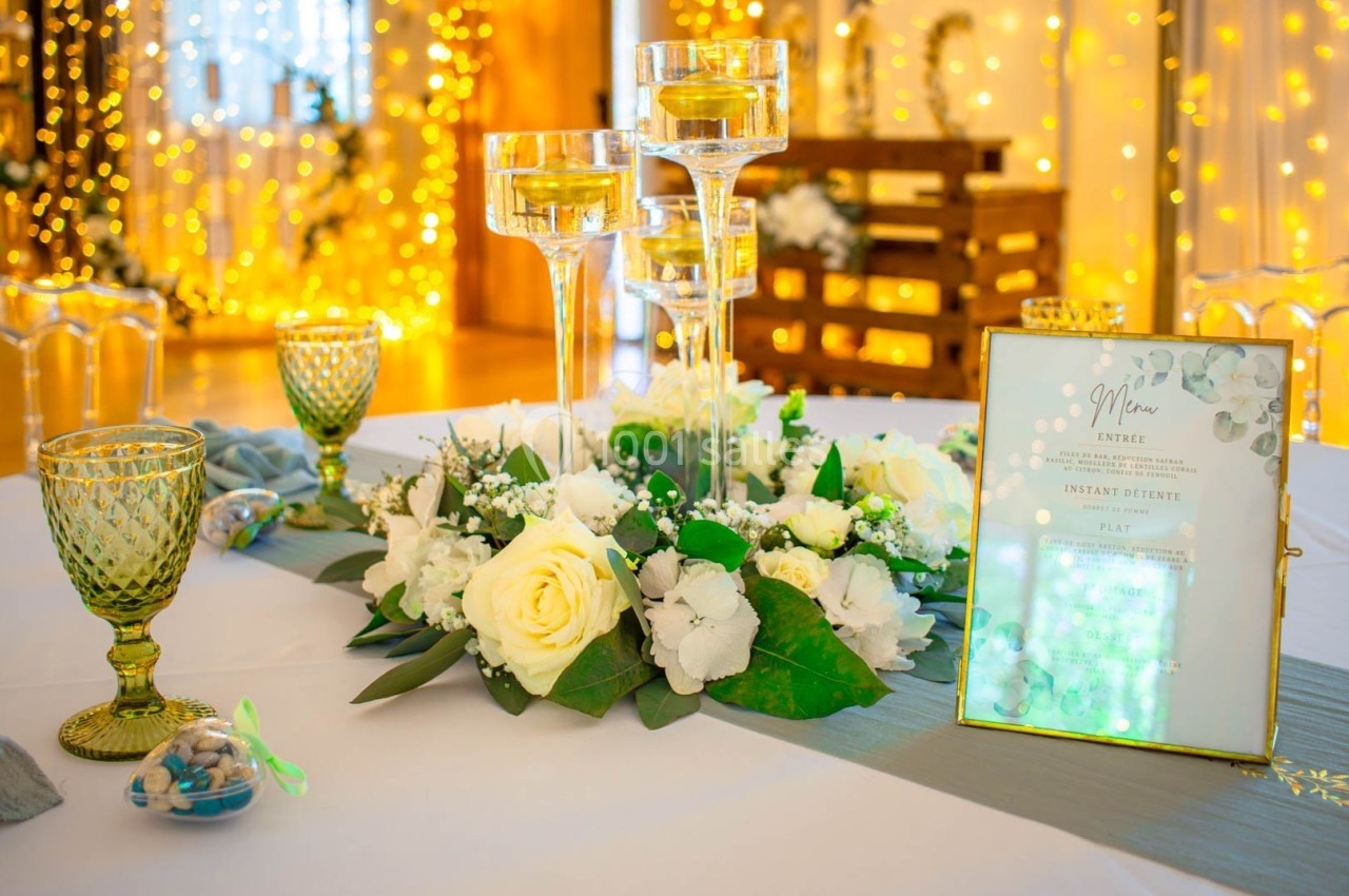 Centre de table avec fleurs blanches, bougies sur supports en verre et menu décoratif, éclairage chaleureux en arrière-plan.