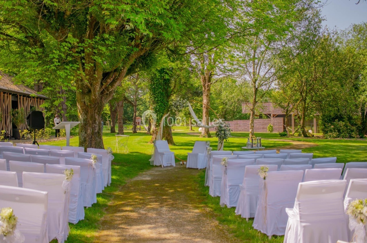 Allée centrale bordée de chaises blanches décorées, menant à un espace de cérémonie en plein air sous des arbres.