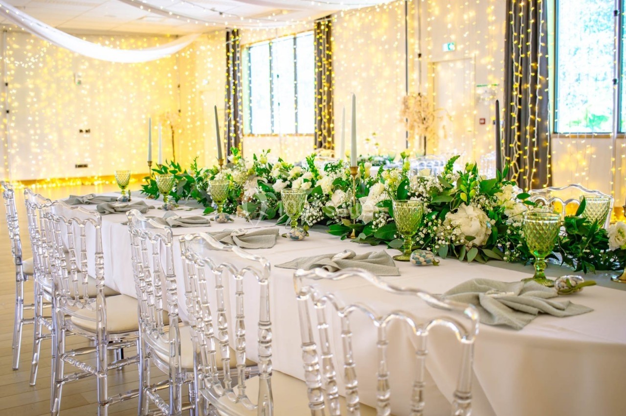 Table de réception élégamment décorée avec nappes blanches, fleurs vertes et blanches, et chaises transparentes.