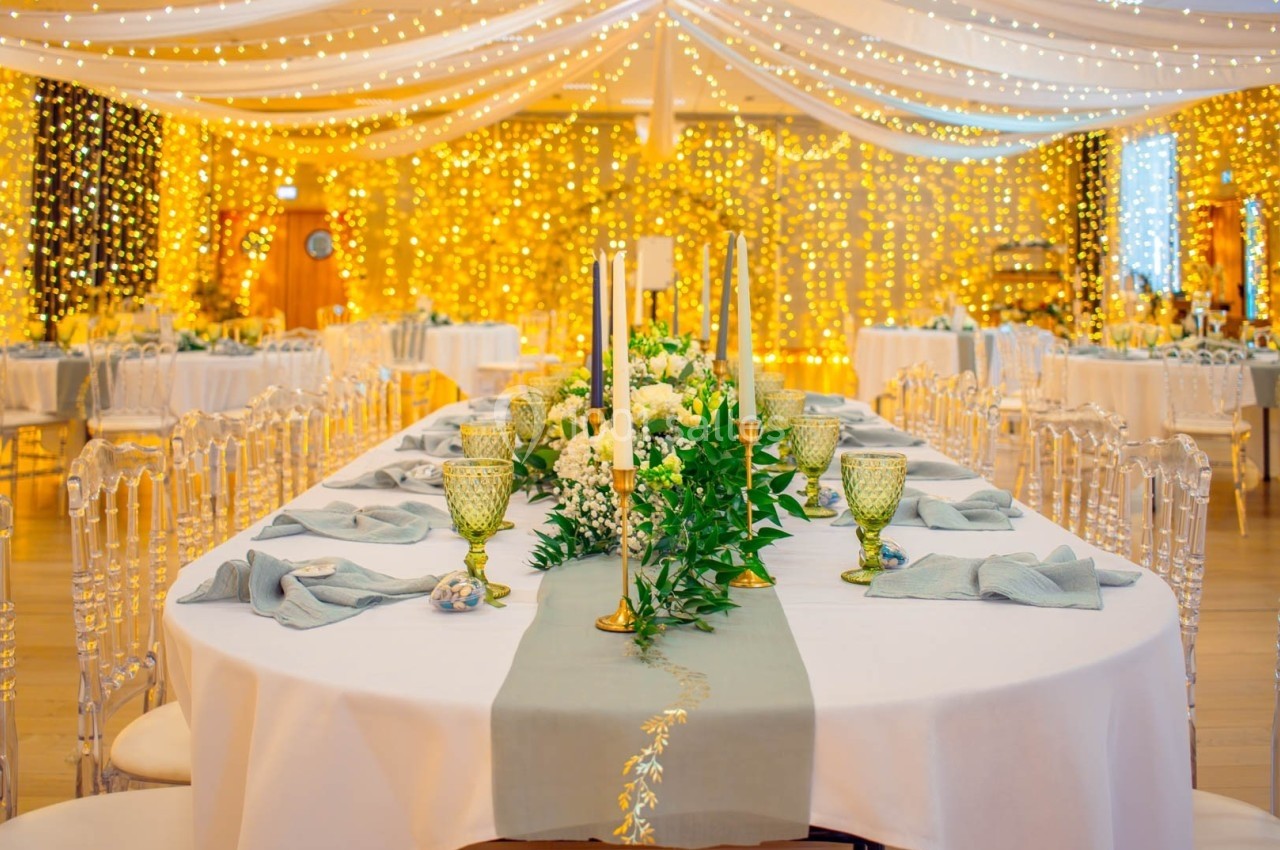 Salle de réception décorée avec des guirlandes lumineuses, tables dressées avec nappes blanches et centres de table fleuris.