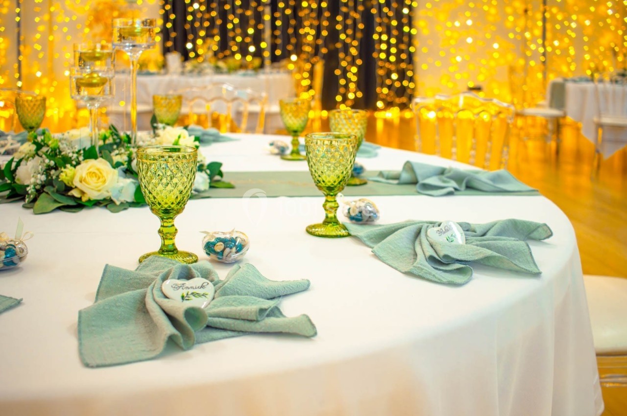 Table décorée pour un événement, avec nappes blanches, serviettes vertes, verres colorés et guirlandes lumineuses en arrière…