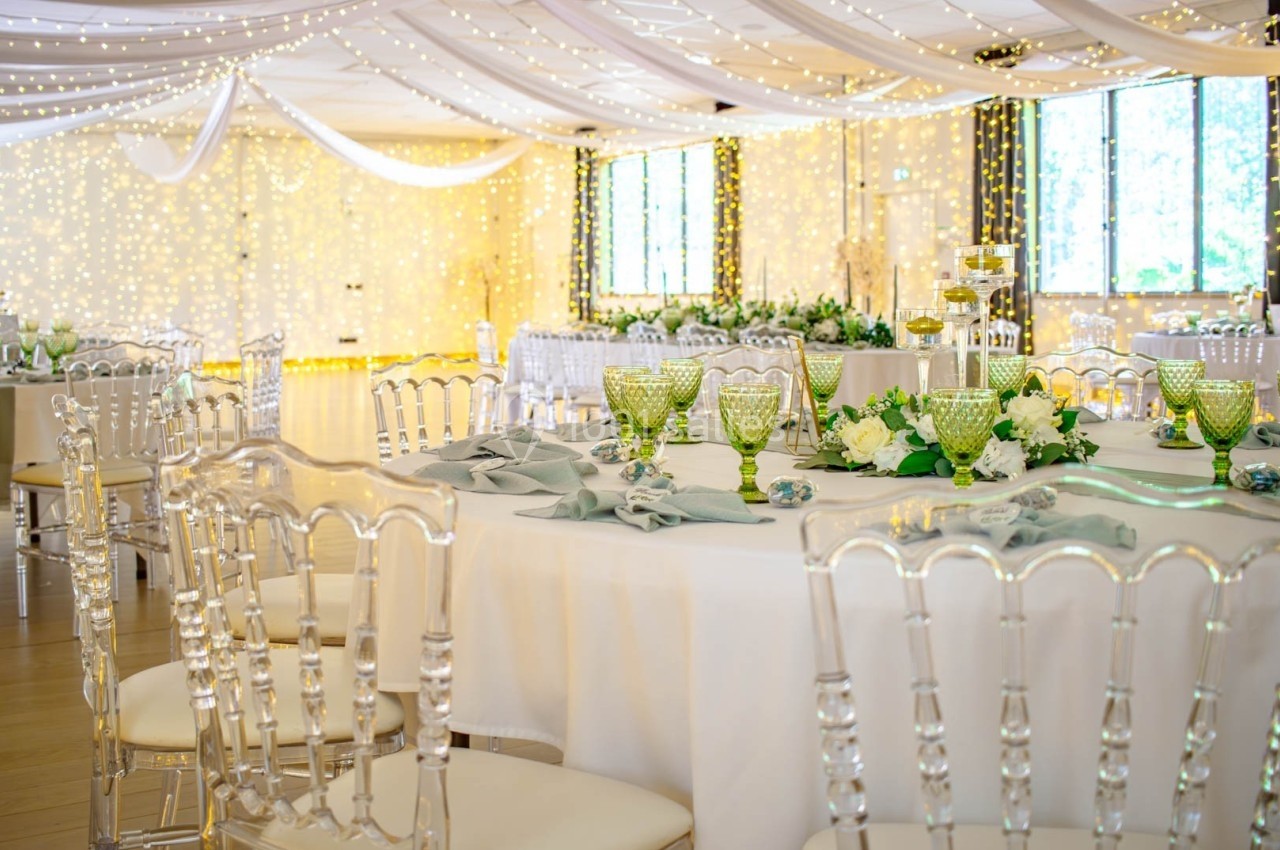 Salle de réception décorée avec des tables élégantes, nappes blanches, chaises transparentes et guirlandes lumineuses…