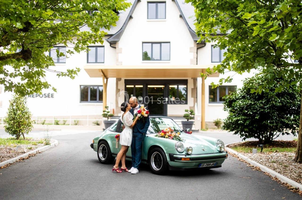 Un couple s'embrasse devant une voiture ancienne verte, garée devant une maison moderne entourée de verdure.
