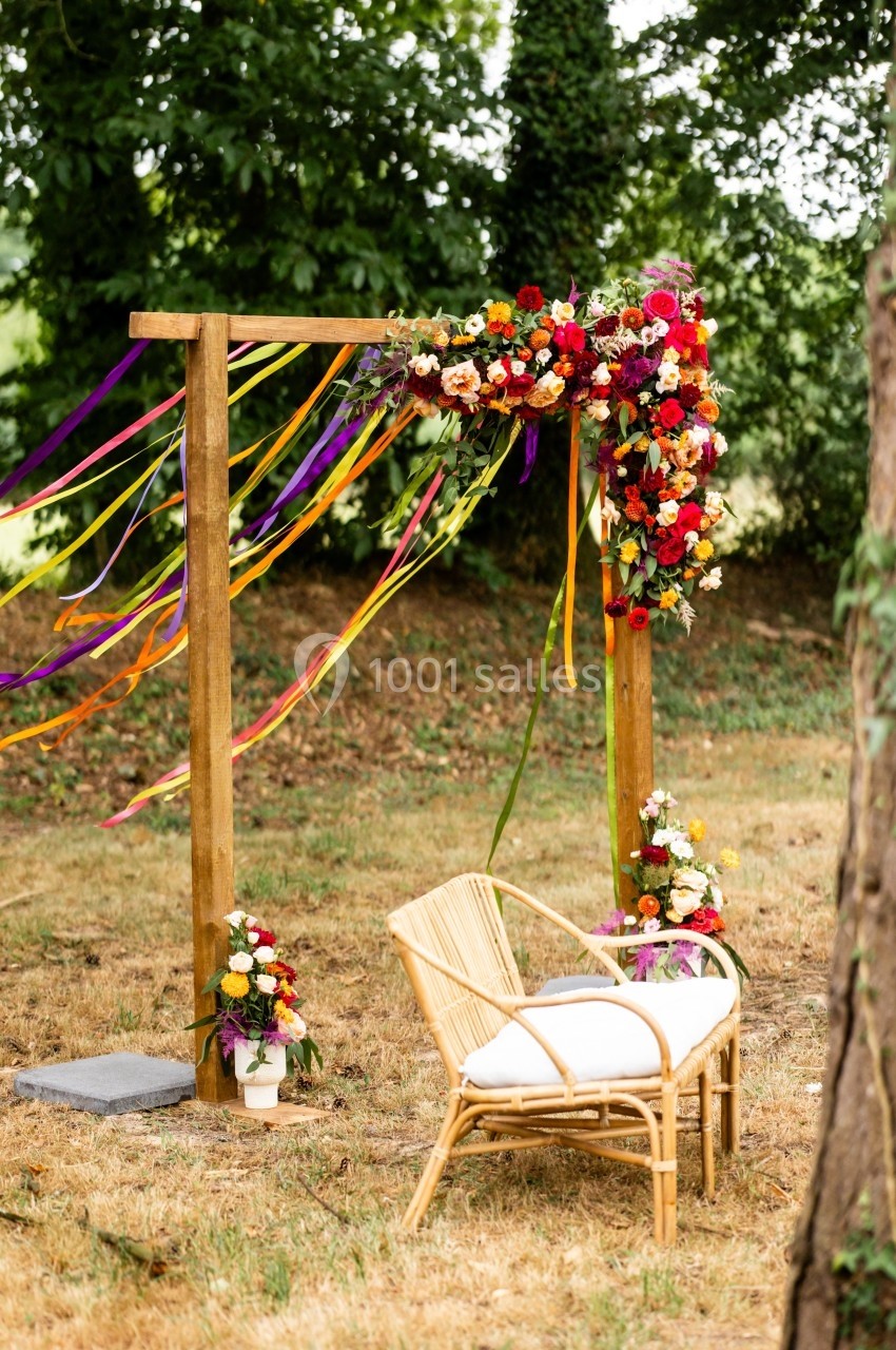 Arche en bois décorée de fleurs colorées et de rubans, avec une chaise en rotin placée sur un terrain herbeux.