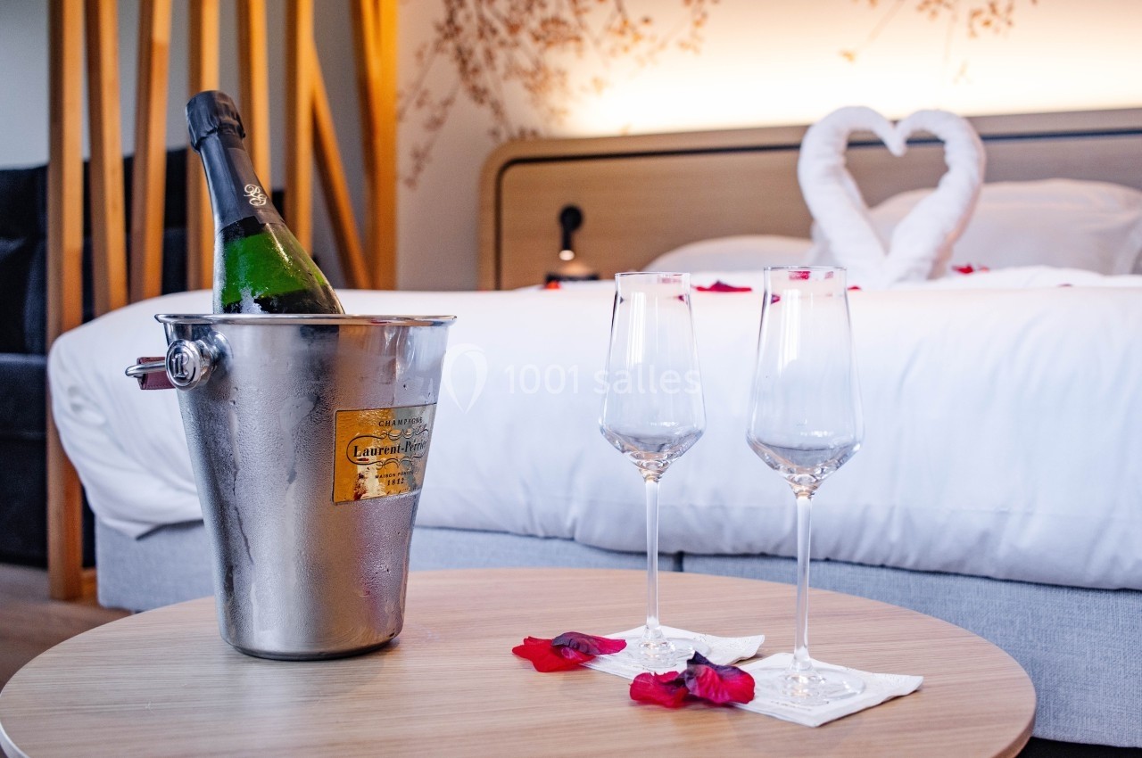 Seau à champagne avec bouteille, deux flûtes et pétales de rose sur une table devant un lit décoré en cœur.