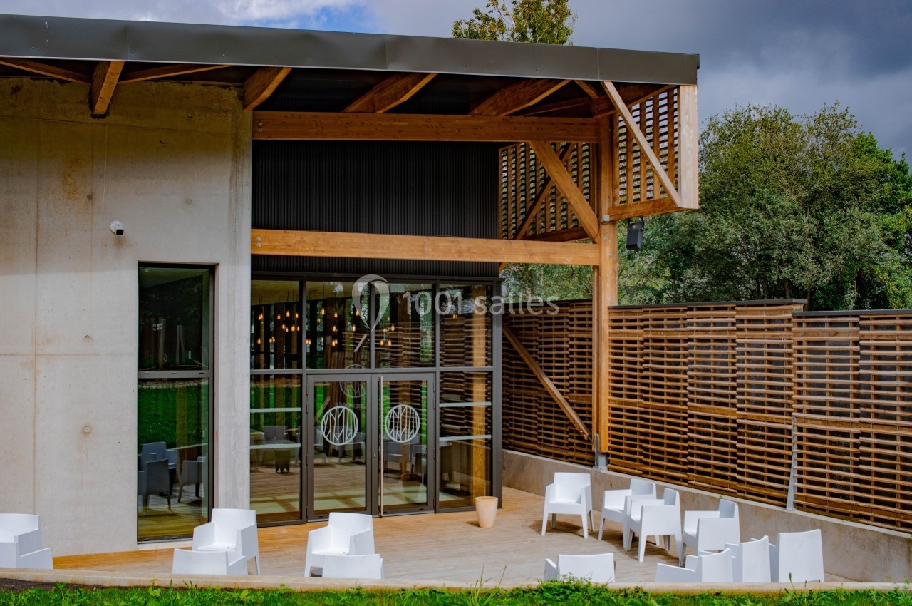 Façade d'un bâtiment moderne avec terrasse en bois, mobilier blanc et grandes baies vitrées.