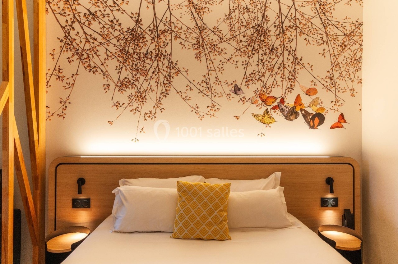 Lit double avec coussin jaune, éclairage doux et décoration murale représentant des branches fleuries et des papillons.