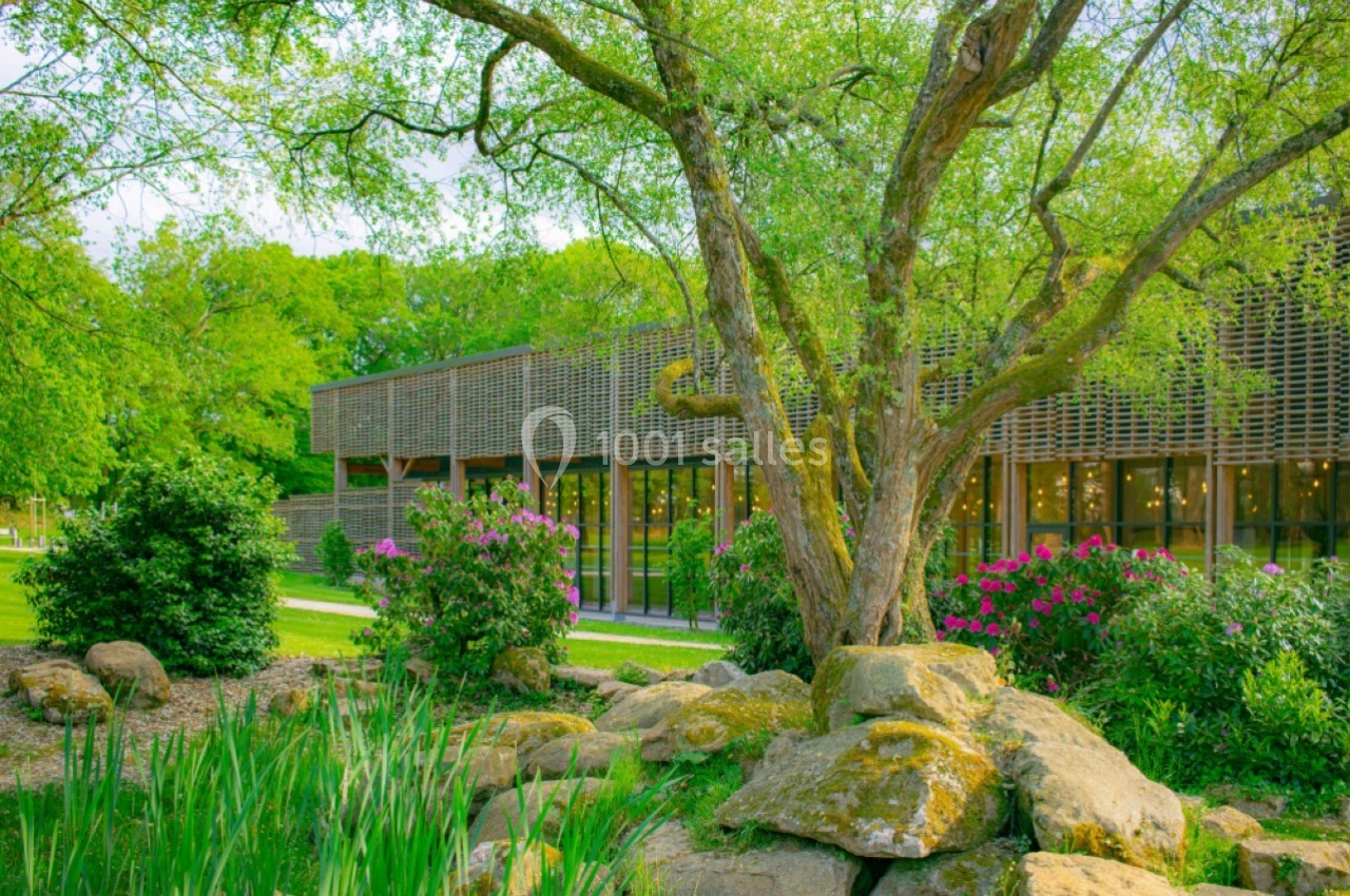 Bâtiment moderne entouré de verdure, avec un arbre au premier plan et des arbustes fleuris.