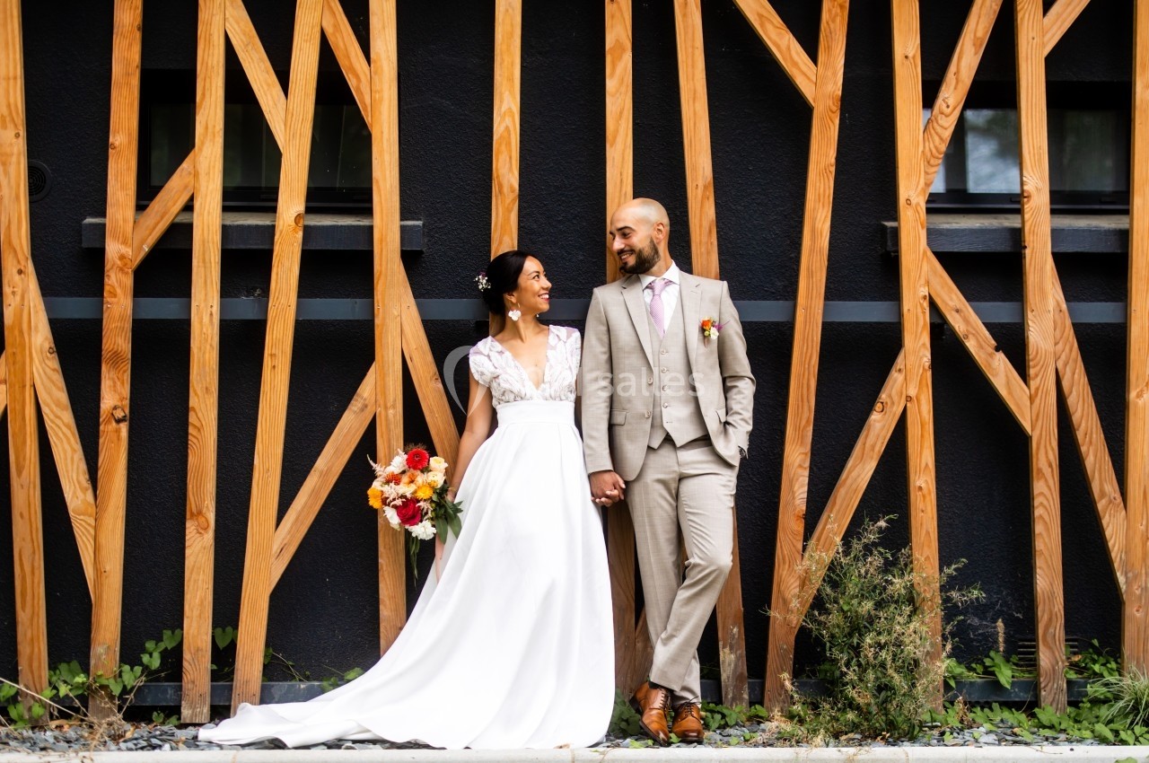 Un couple de mariés sourit en se tenant la main devant une structure en bois moderne.