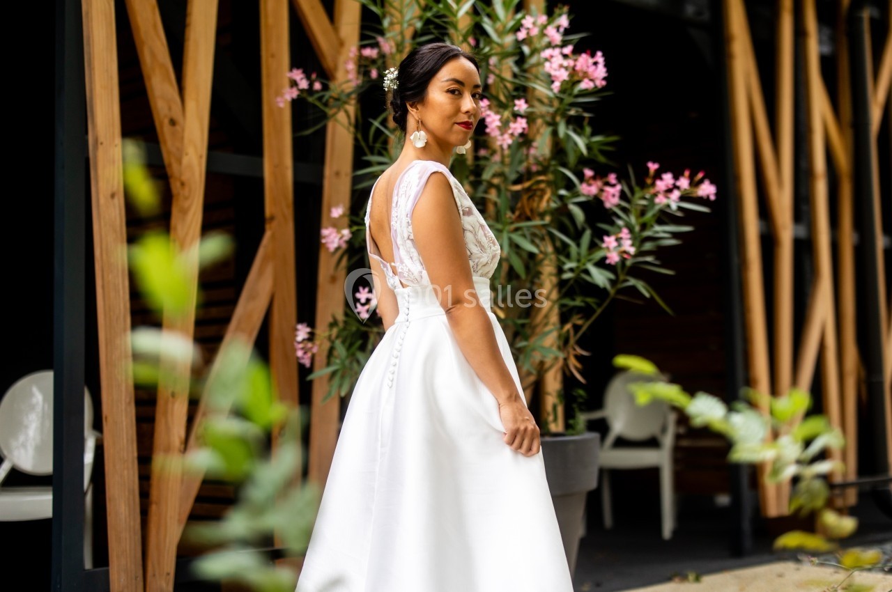 Femme en robe de mariée blanche, de profil, devant des plantes et une structure en bois.