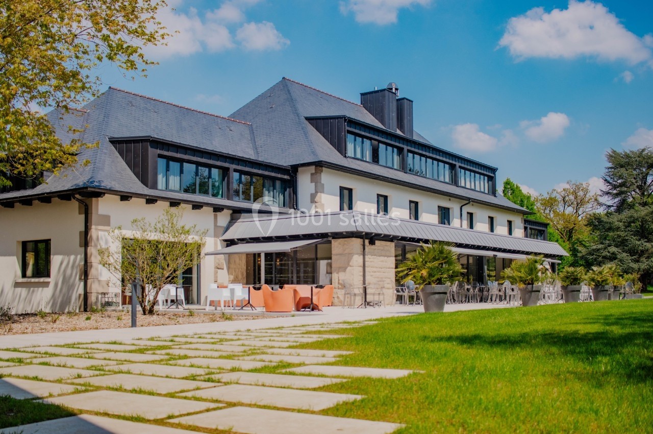 Bâtiment moderne avec toit en ardoise, terrasse aménagée et jardin verdoyant sous un ciel bleu.