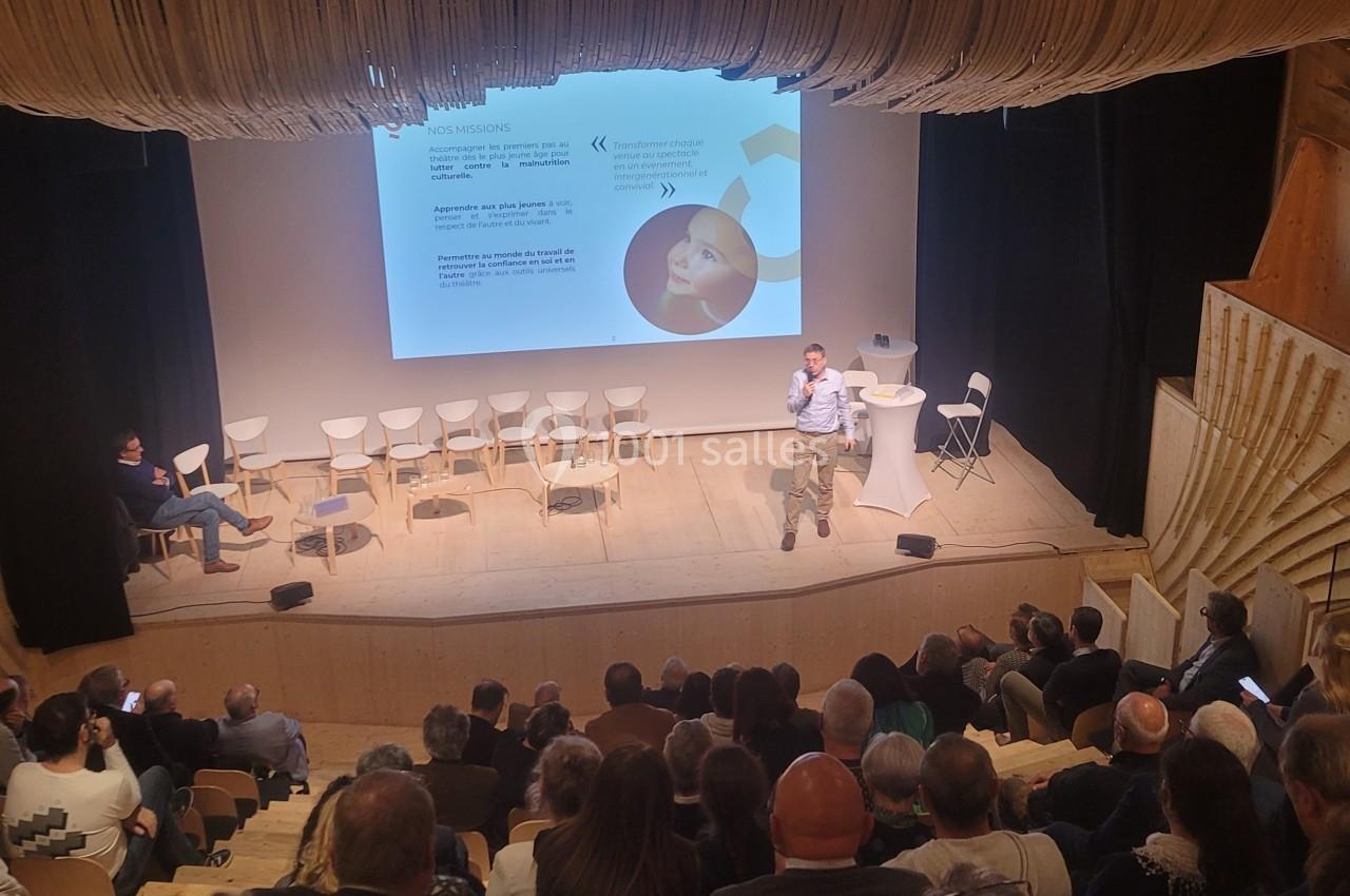 Un conférencier s'adresse à un public dans un amphithéâtre en bois, avec une présentation projetée à l'écran.