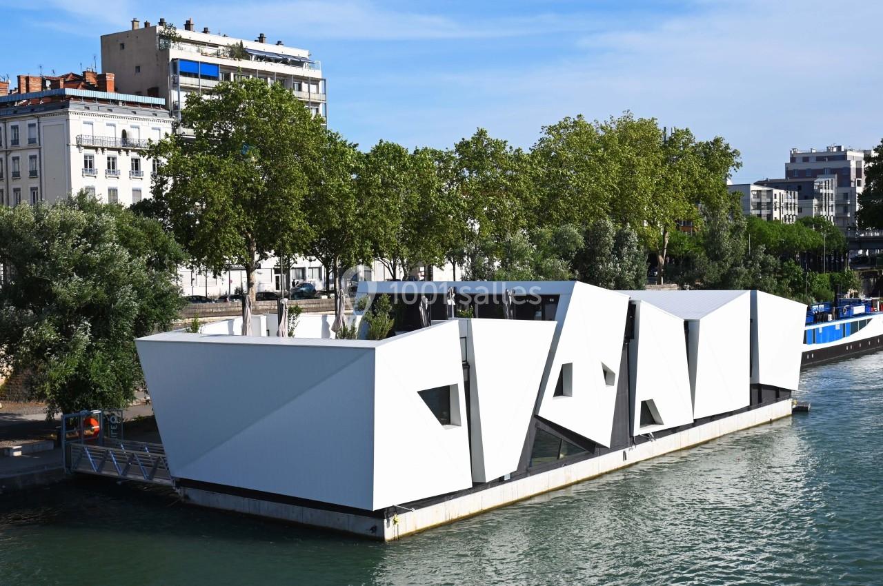 Bâtiment flottant moderne en structure blanche géométrique, amarré sur une rivière bordée d'arbres et d'immeubles.