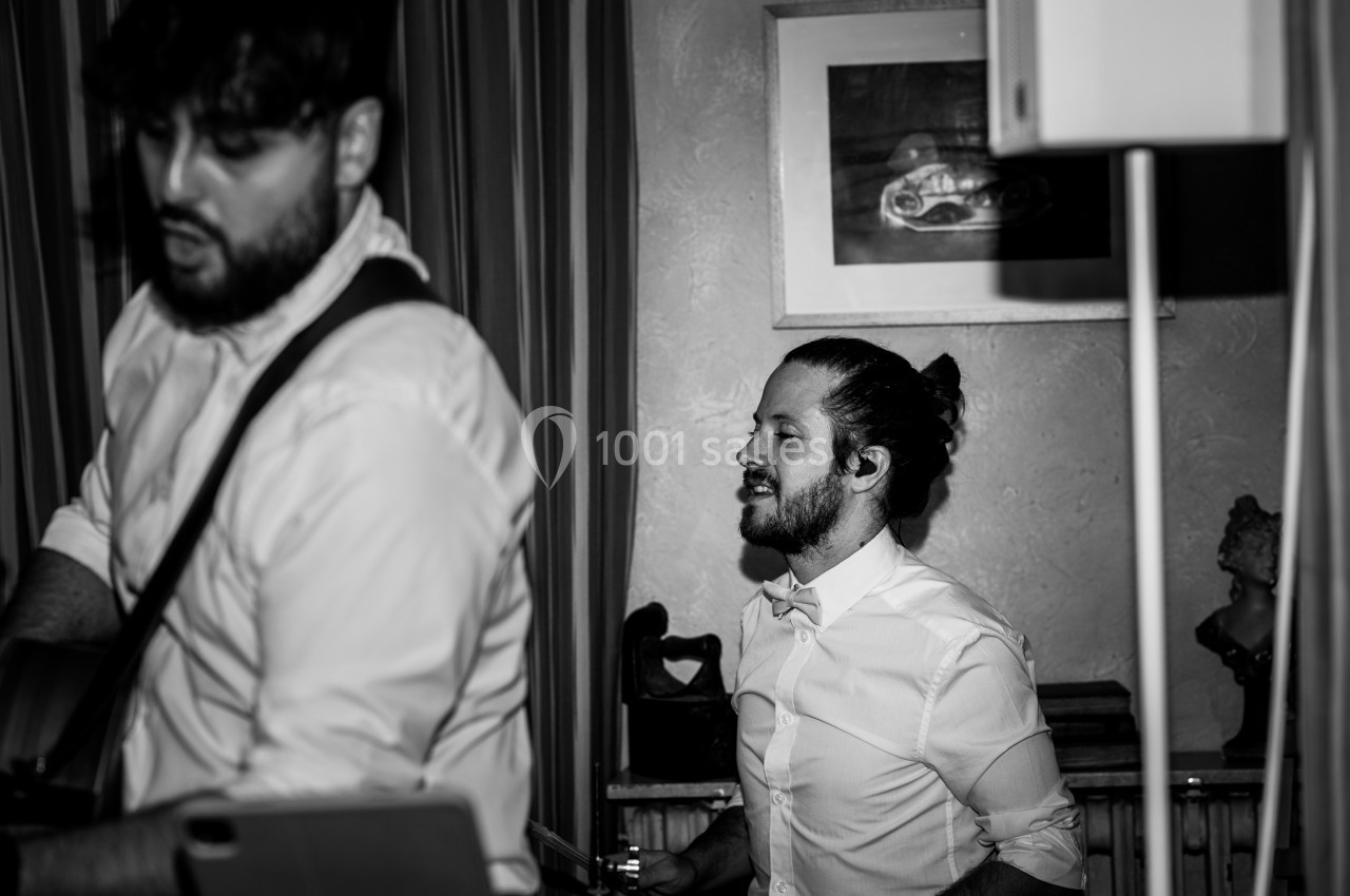 Deux musiciens en chemise blanche jouent dans une pièce intérieure, l'un à la guitare et l'autre à la batterie.