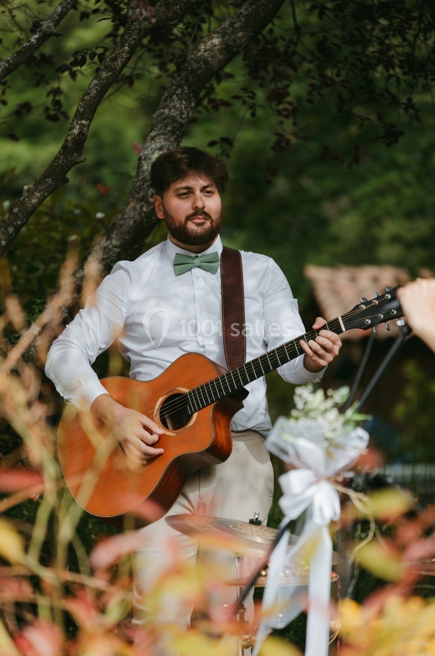 Un homme joue de la guitare acoustique en extérieur, entouré de verdure et de décorations florales.