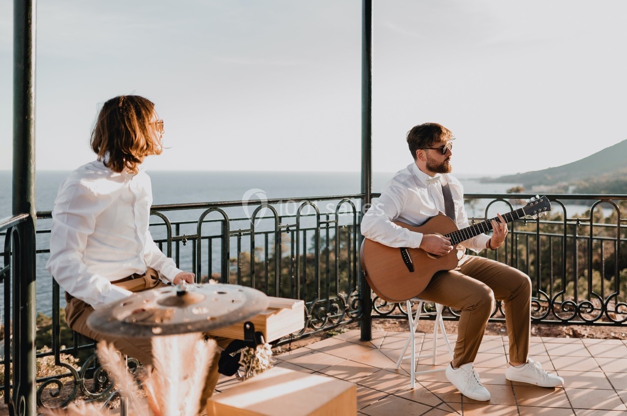 Deux musiciens jouent de la guitare et des percussions sur une terrasse surplombant la mer par temps ensoleillé.