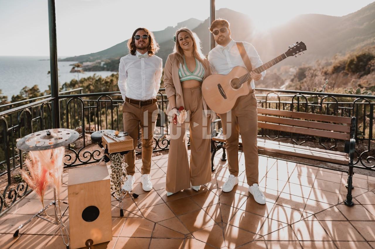 Trois musiciens posent sur une terrasse ensoleillée avec vue sur la mer et les montagnes, guitare et percussions visibles.