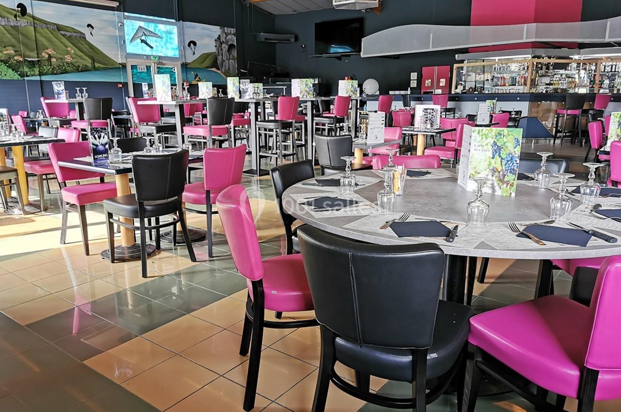 Salle de restaurant lumineuse avec des tables dressées, chaises roses et noires, et un bar en arrière-plan.