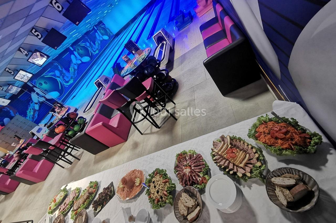 Buffet varié avec charcuterie et légumes en premier plan, dans un bowling avec pistes et sièges colorés en arrière-plan.