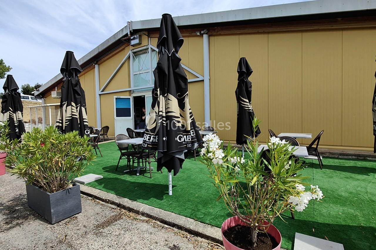 Terrasse extérieure avec tables, chaises et parasols noirs, entourée de plantes en pots, devant un bâtiment jaune.