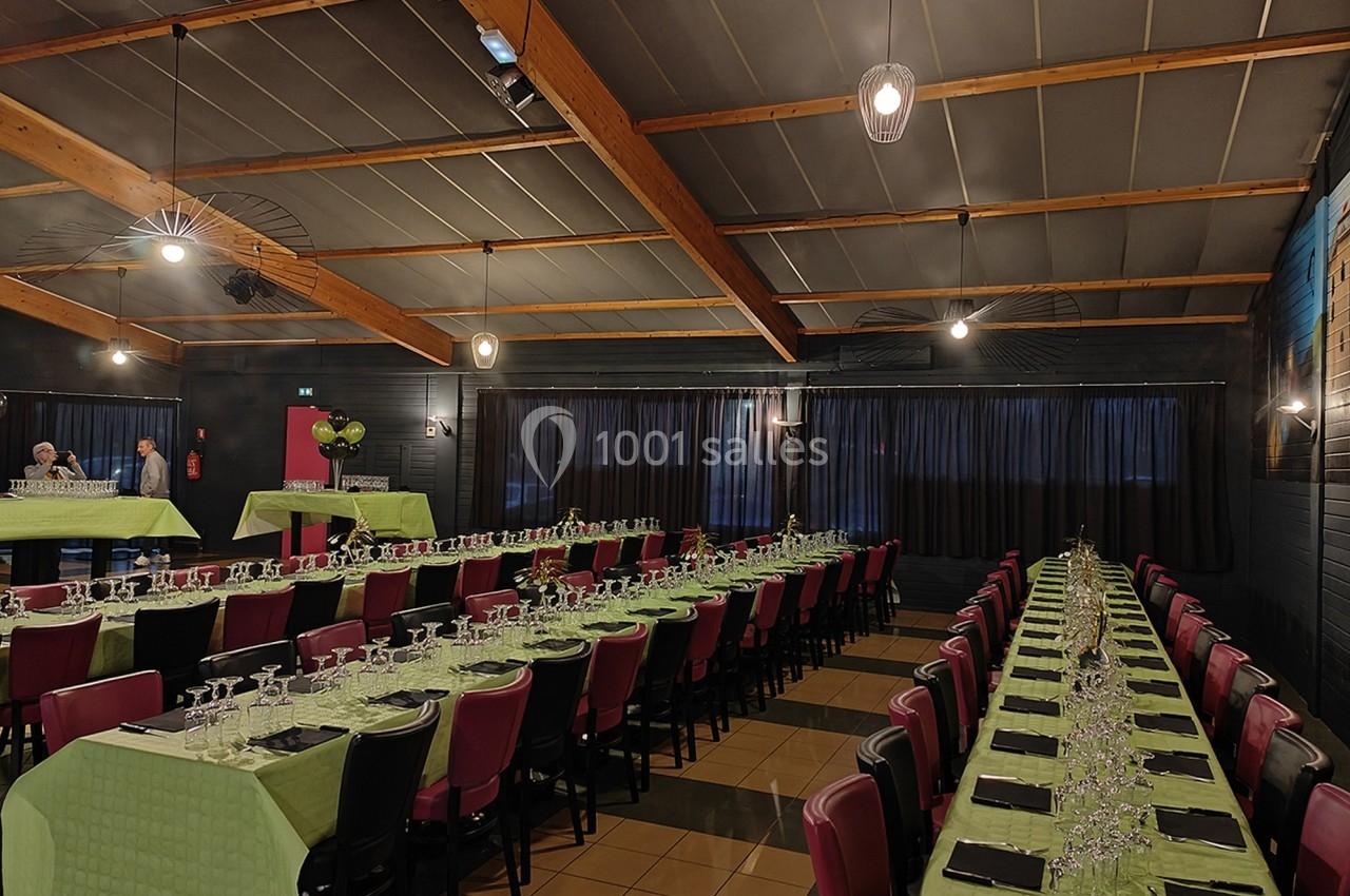Salle de réception avec de longues tables dressées, nappes vertes, chaises rouges et éclairage tamisé.