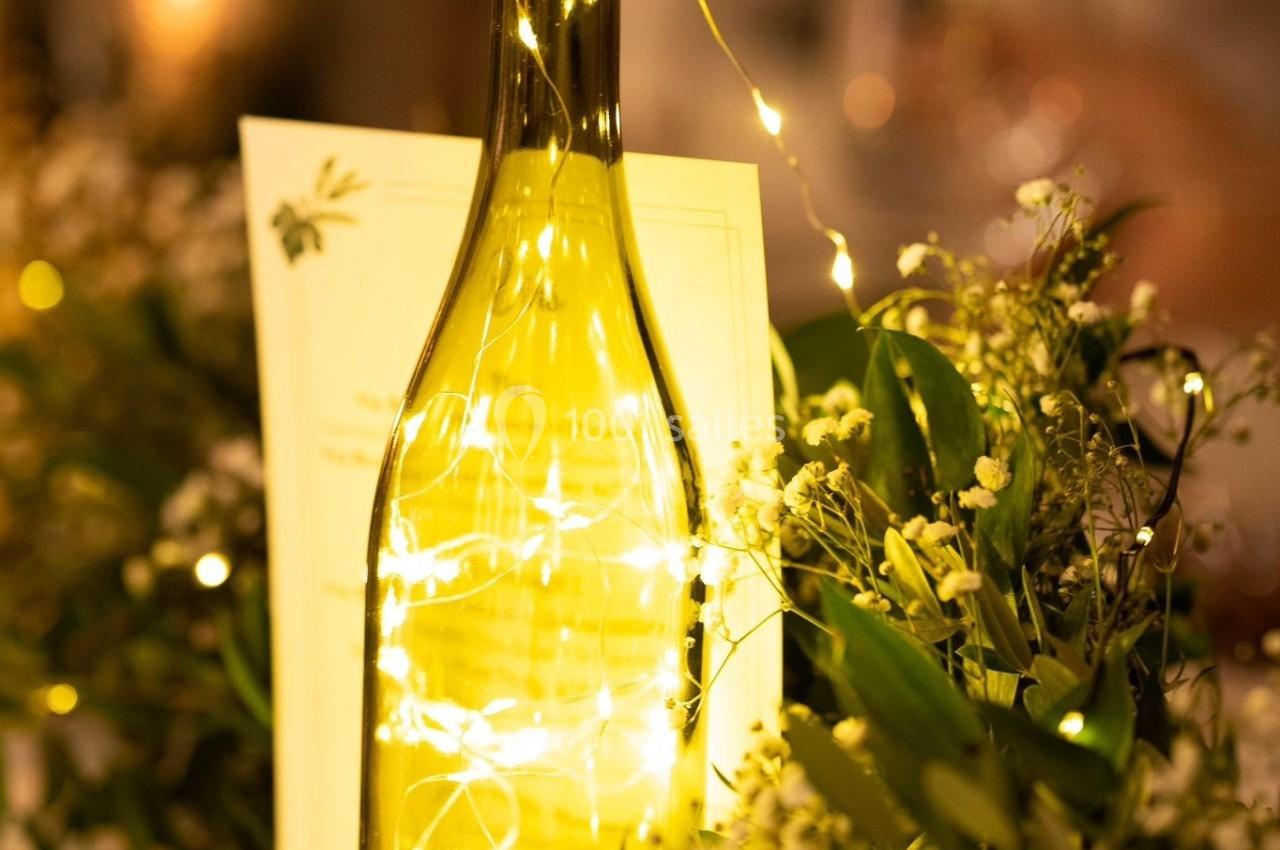Bouteille en verre éclairée par une guirlande lumineuse, entourée de feuillage et de fleurs sur une table décorée.