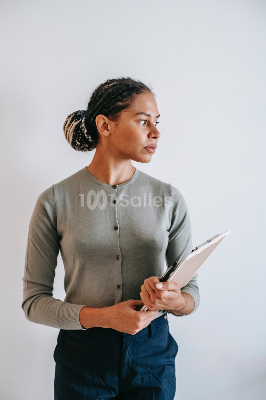 Femme debout tenant un clipboard, regardant sur le côté, vêtue d'un cardigan gris et d'un pantalon foncé.