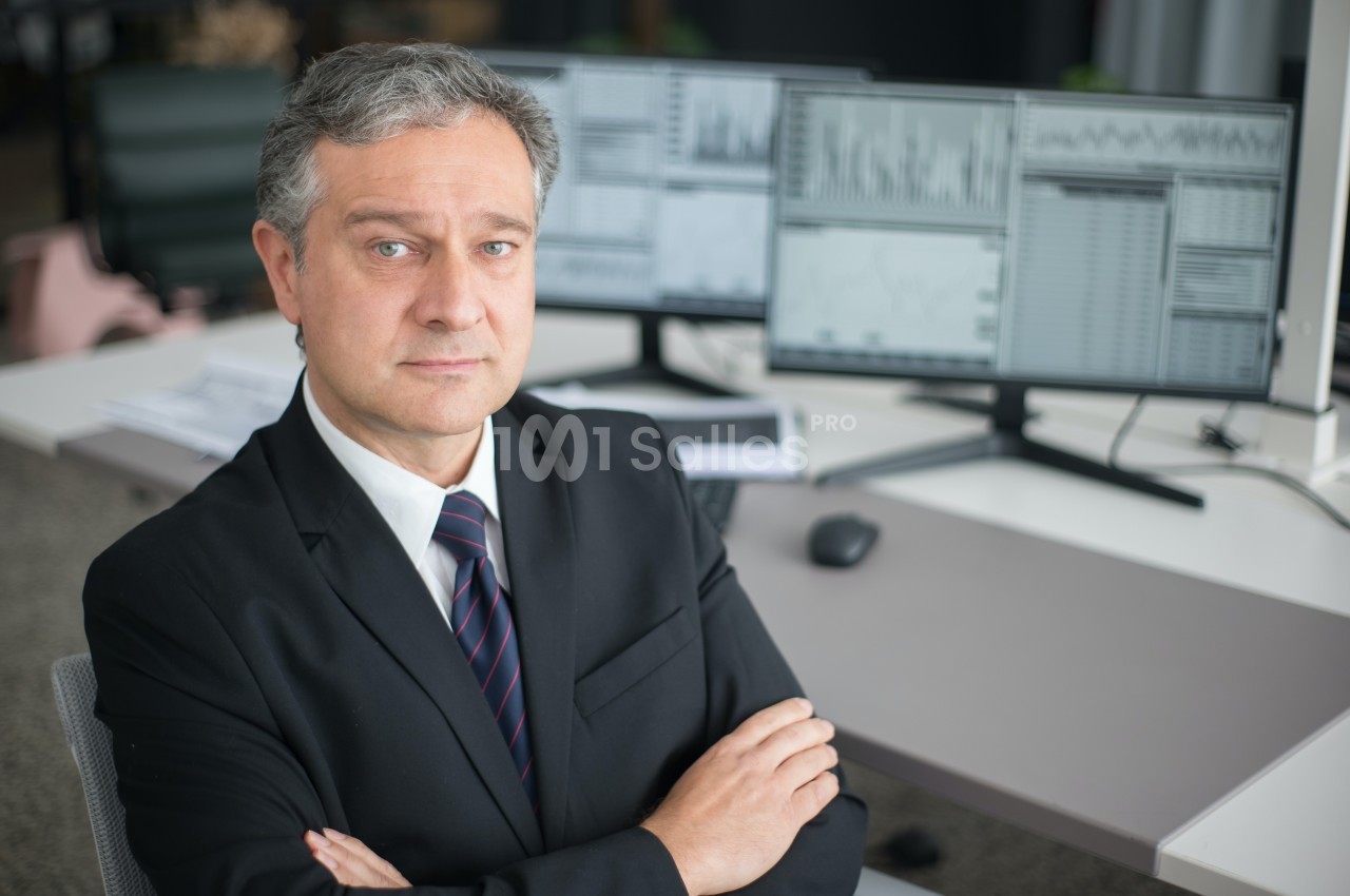 Homme en costume assis devant un bureau avec plusieurs écrans affichant des graphiques et des données.