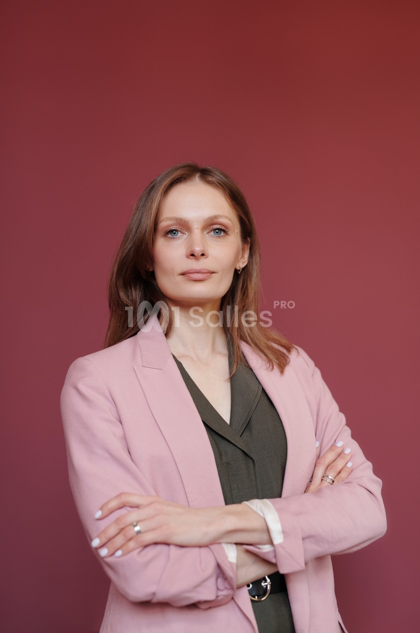 Femme aux cheveux châtains portant un blazer rose et une chemise kaki, posant devant un fond rose uni.
