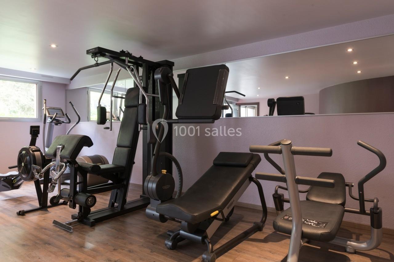 Salle de sport équipée de machines de musculation et de cardio, avec un grand miroir et un sol en bois.
