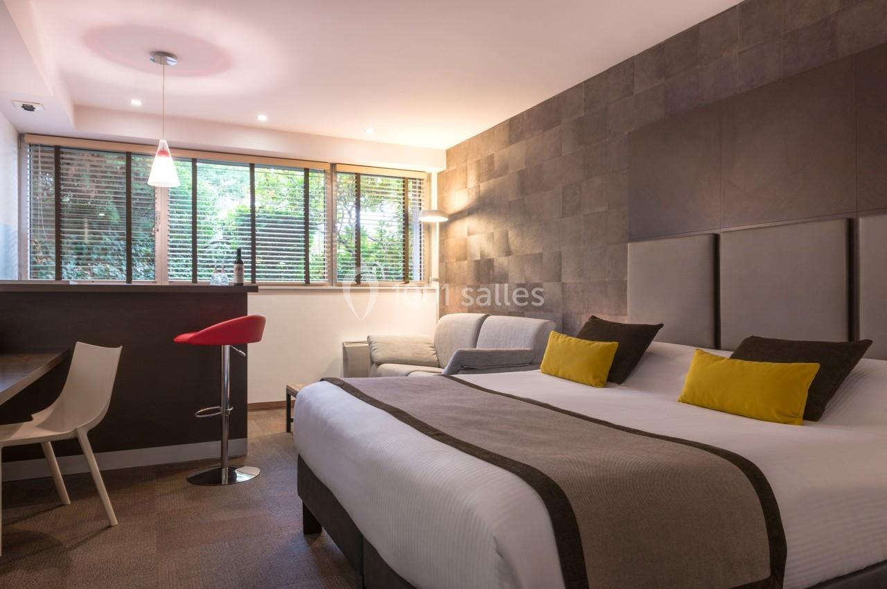 Chambre moderne avec lit double, fauteuil, bureau, tabouret rouge et grande fenêtre donnant sur un espace vert.