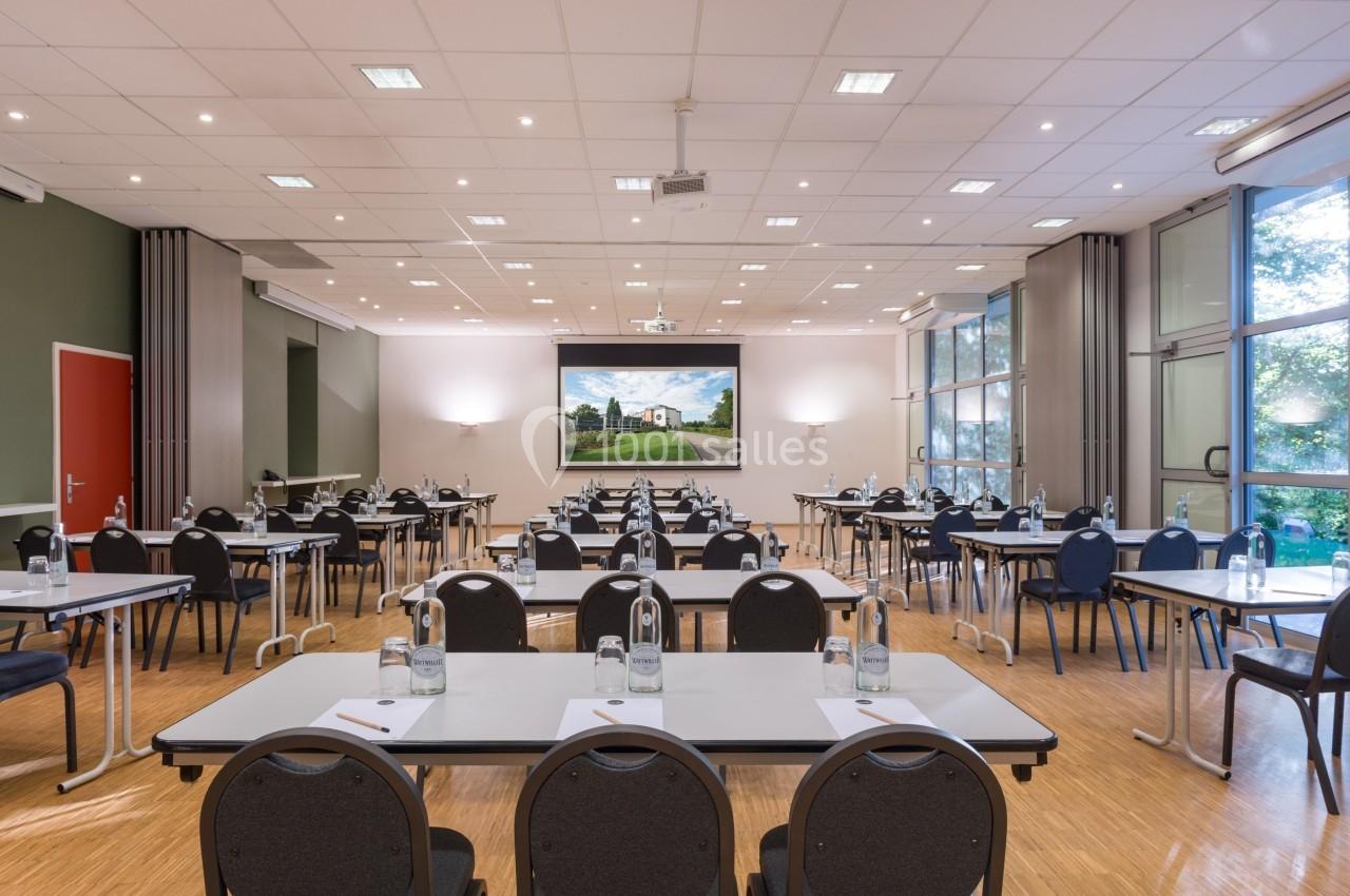 Salle de conférence lumineuse avec des tables et chaises alignées, écran de projection allumé en fond.